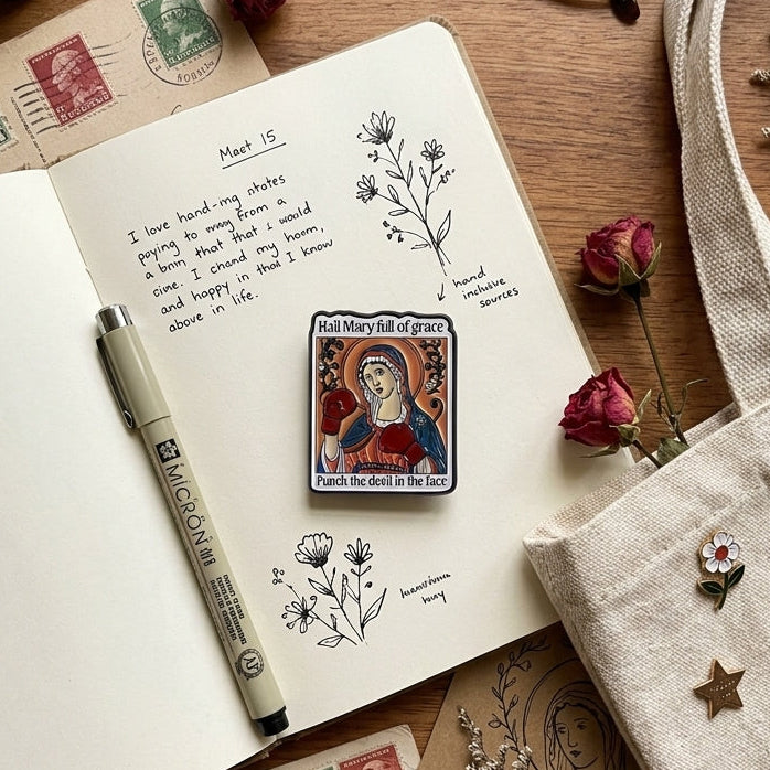 A styled flat-lay featuring a rectangular religious pin with a depiction of Mary, which reads 'Hail Mary full of grace, Punch the devel in the face', pinned to an open notebook. The scene is decorated with a beige canvas tote bag, eyeglasses, dried roses, stamps, a pen, and eucalyptus on a wooden tabletop.