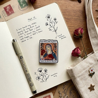A styled flat-lay featuring a rectangular religious pin with a depiction of Mary, which reads 'Hail Mary full of grace, Punch the devel in the face', pinned to an open notebook. The scene is decorated with a beige canvas tote bag, eyeglasses, dried roses, stamps, a pen, and eucalyptus on a wooden tabletop.