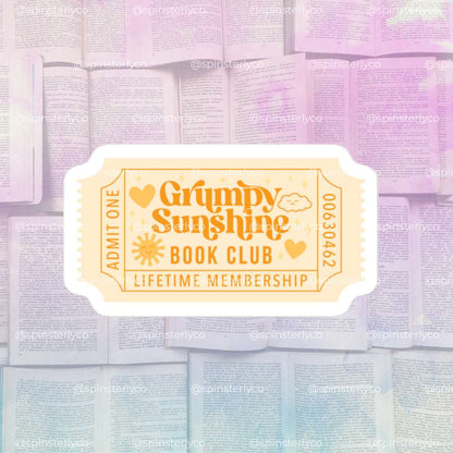 A retro-style ticket graphic reading 'Grumpy Sunshine Book Club Lifetime Membership' with heart icons and a sun, centered over a pastel background of open books.