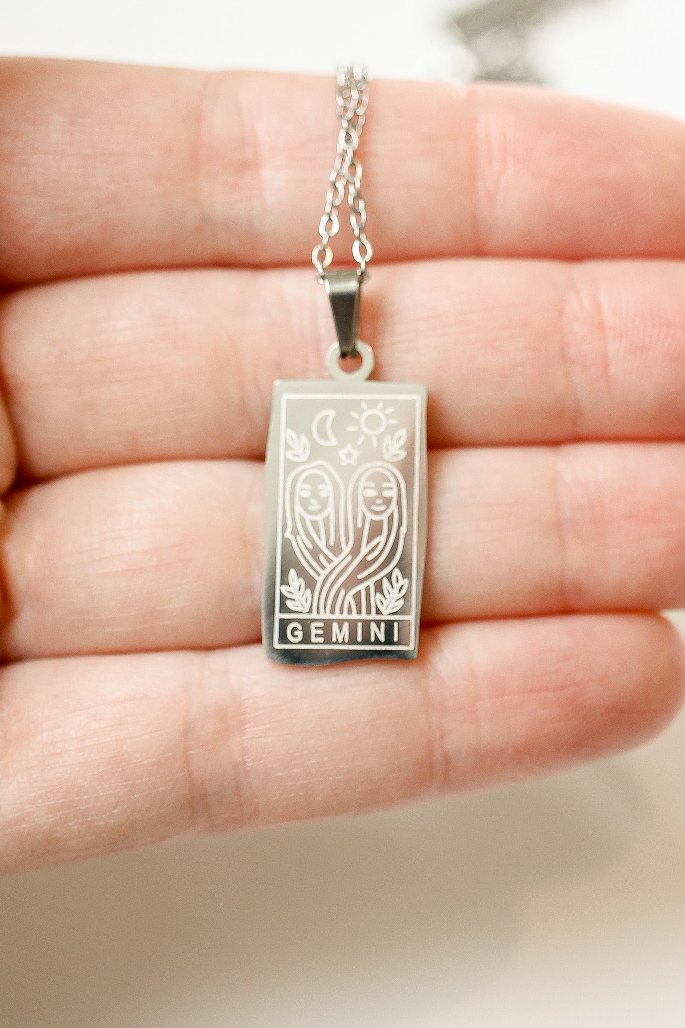 Silver 'Gemini' necklace pendant held in a hand against a neutral background