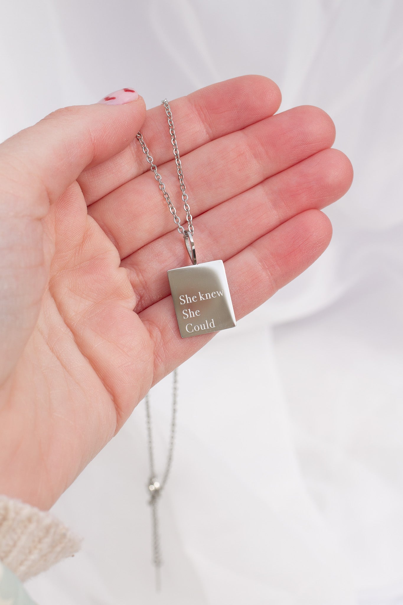 Hand holding a silver necklace with a pendant that reads 'She knew she could'.