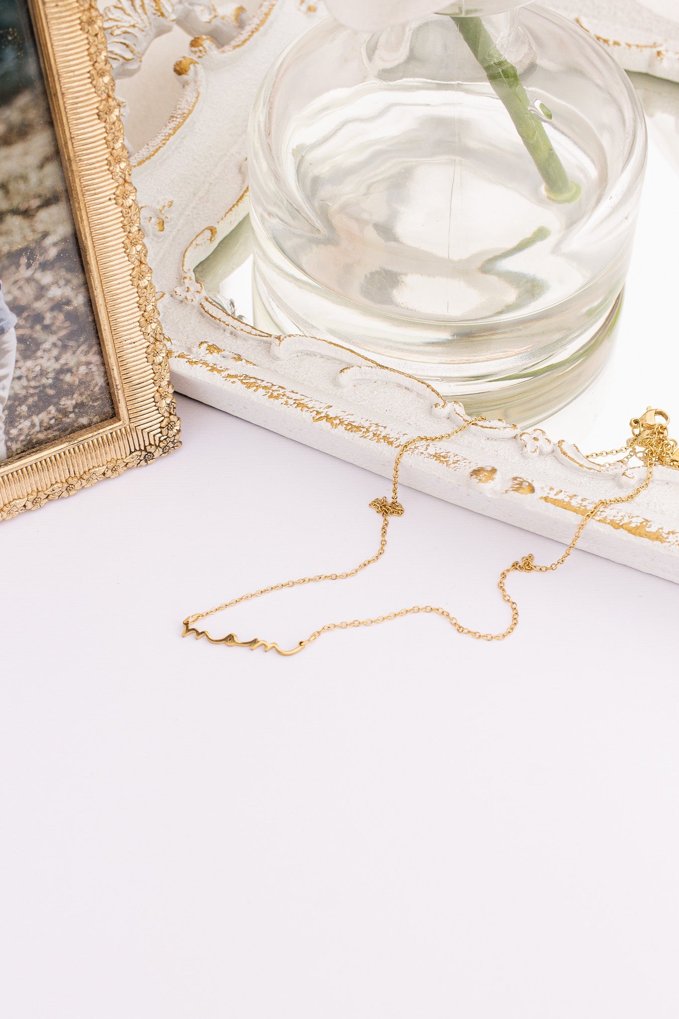 Gold necklace on a white surface with a glass of water and decorative frame in the background