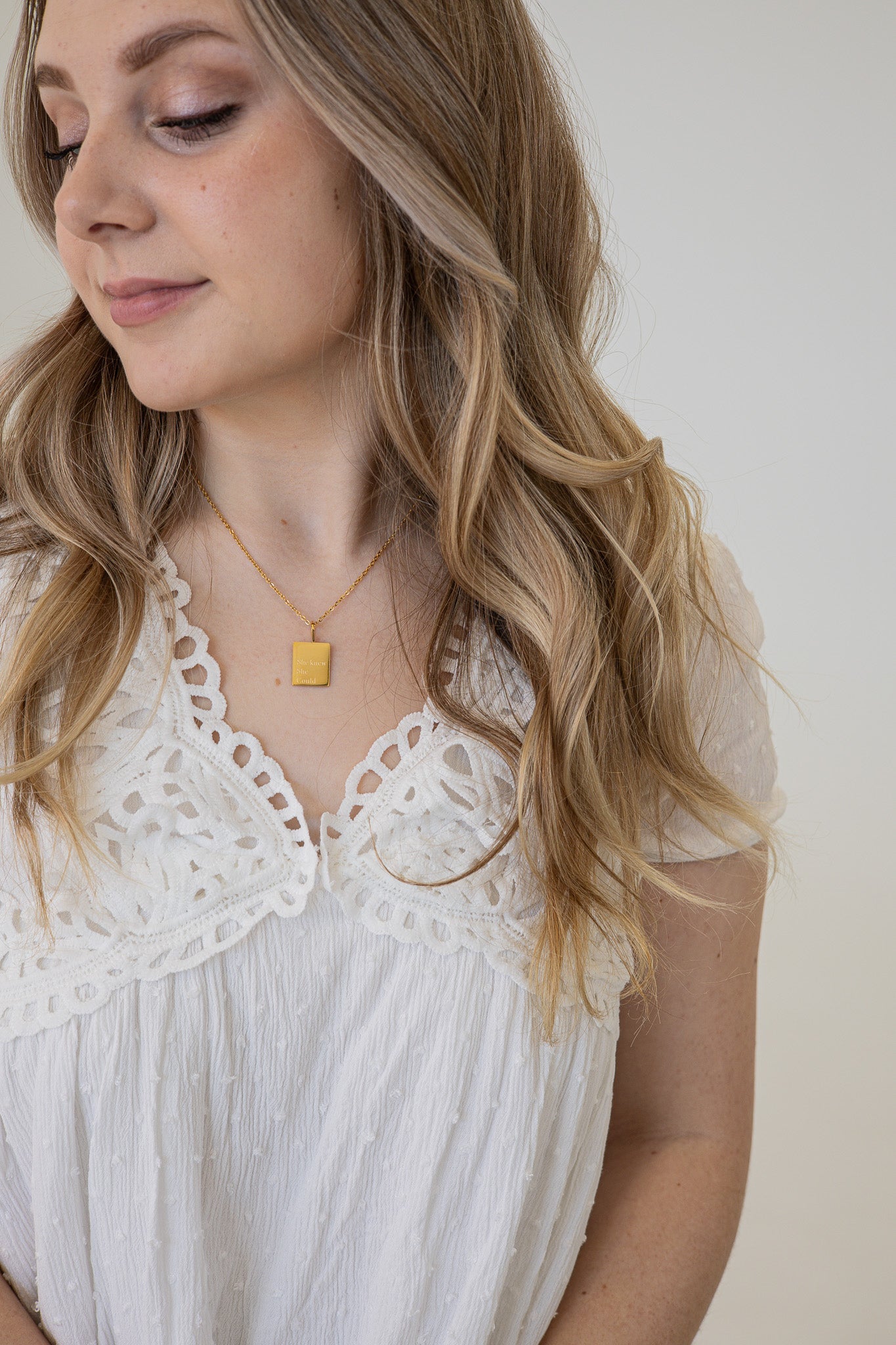 Woman wearing a white lace top and gold necklace with a small pendant against a plain background