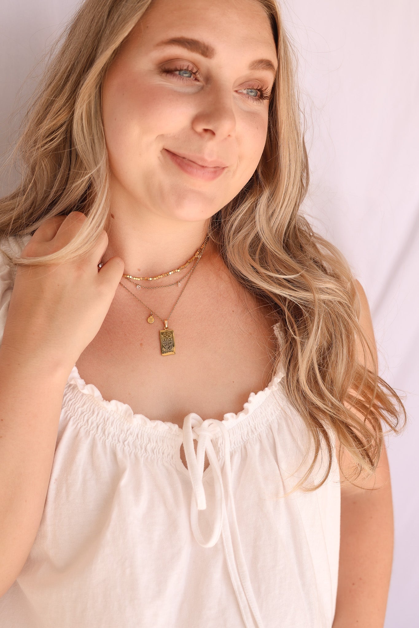 A woman with blonde hair wearing a white ruffled top and layered gold necklaces, one with a small green rectangular pendant, against a light background.