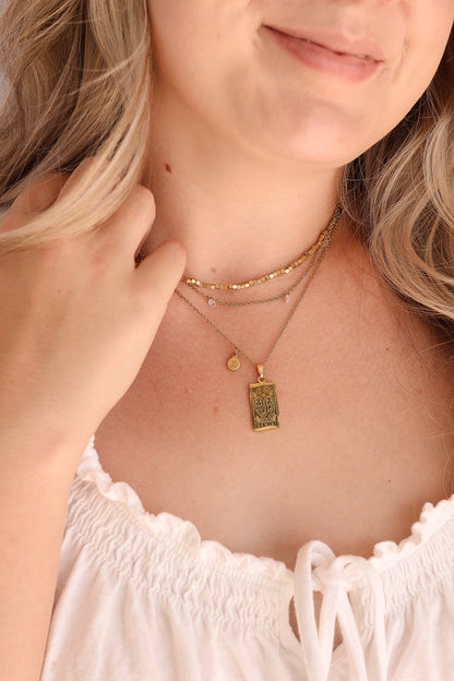 A close-up of a woman wearing layered gold necklaces, including a textured rectangular pendant, a delicate chain with a small round charm, and a thin chain with tiny clear stones, paired with a white ruffled top.
