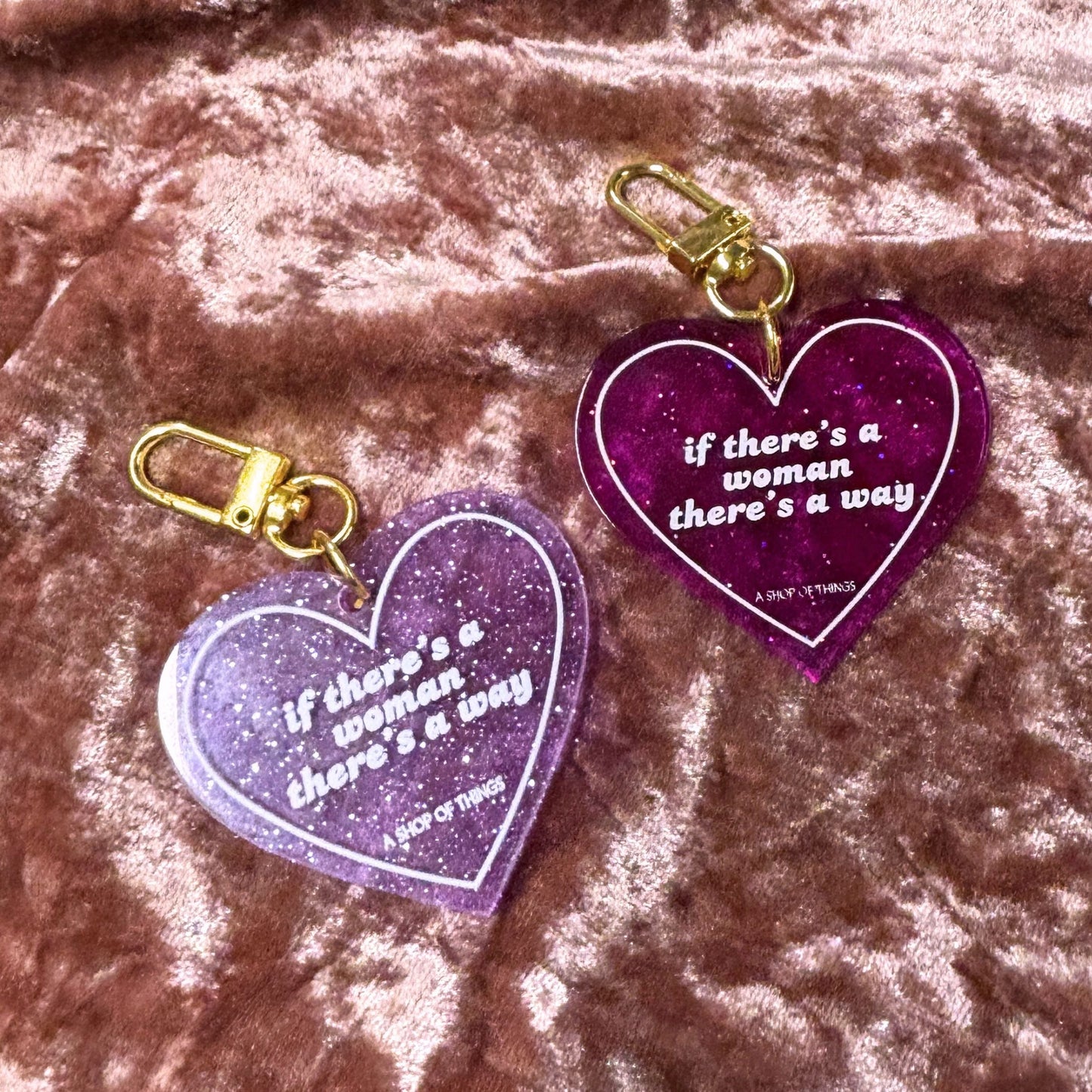 Two purple heart-shaped keychains with gold clips on a pink textured background.