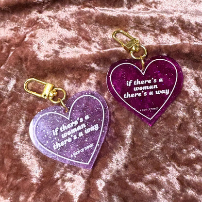 Two purple heart-shaped keychains with gold clips on a pink textured background.