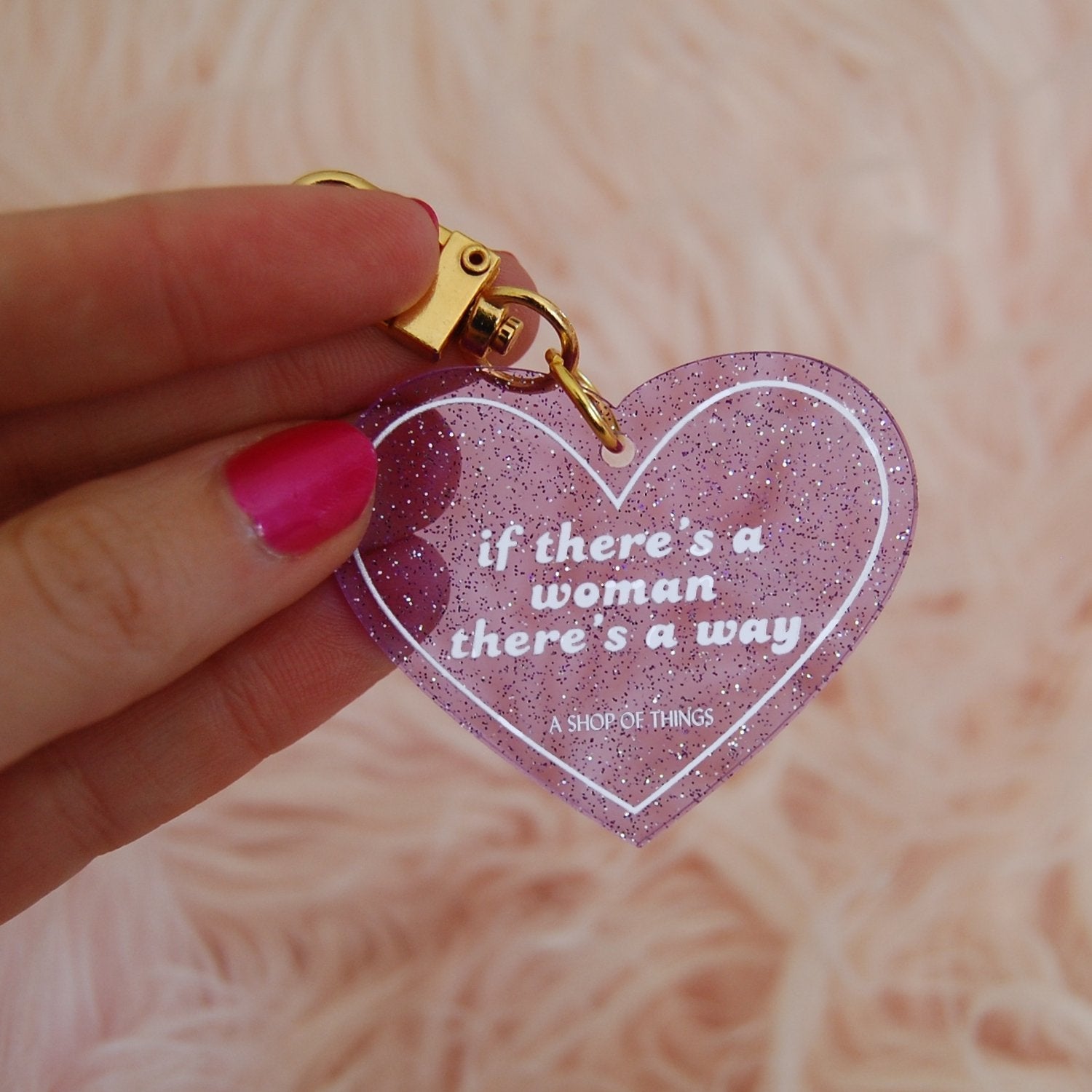 Heart-shaped keychain with motivational quote held by a hand against a soft pink background