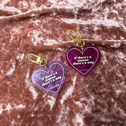 Two purple heart-shaped keychains with gold clasp on a pink textured fabric background.