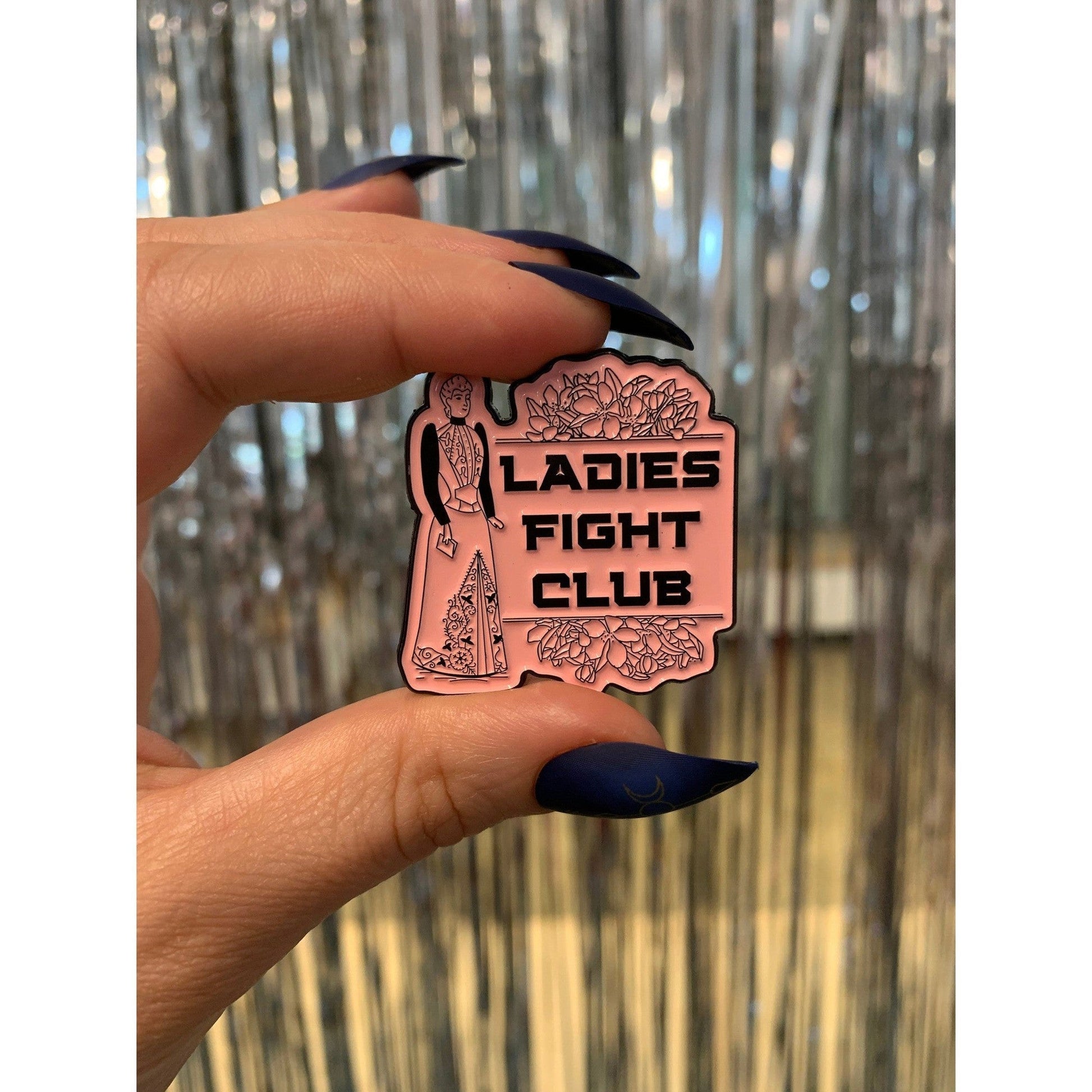Copper 'Ladies Fight Club' pin held between fingers against a reflective surface.