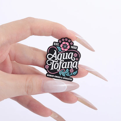 A hand with long manicured nails holds a small decorative pin that reads 'Aqua Tofana' with floral details on a plain white background.
