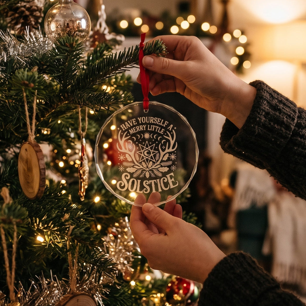 A person's hands hang a round clear glass Christmas ornament with engraved text and a red ribbon from a decorated Christmas tree in a cozy, warmly lit living room.