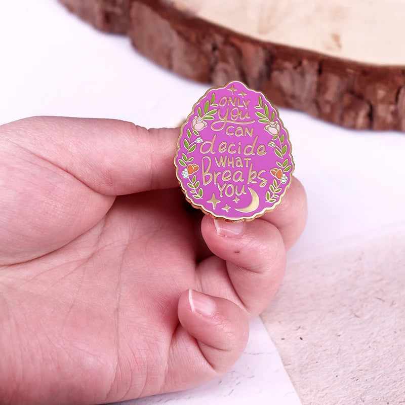 A hand holding a pink enamel pin with gold lettering that reads 'Only You Can decide What Breaks You' surrounded by floral and crescent moon details.