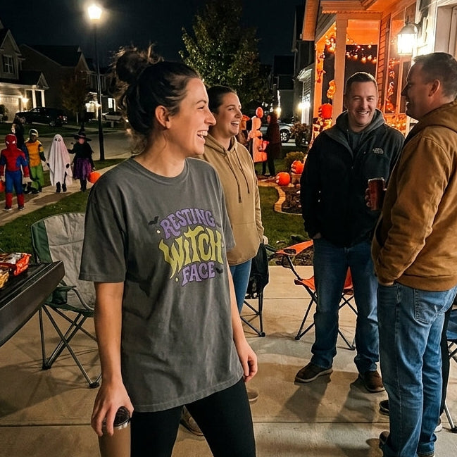 A woman wearing a gray graphic t-shirt with 'RESTING WITCH FACE' in yellow and purple text, black leggings, and brown boots stands on a front porch at night. Other people are gathered nearby on folding chairs and around Halloween decorations.