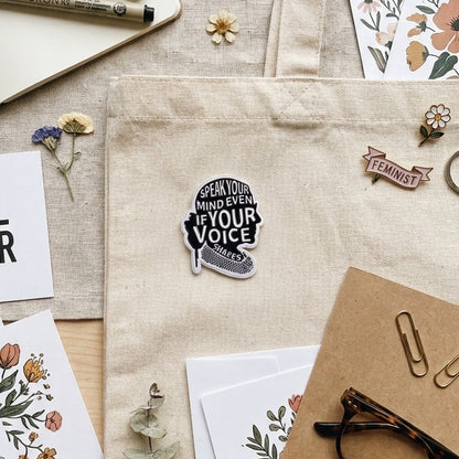 A styled flat-lay featuring a beige canvas tote bag with several decorative pins attached, including a silouhette of Ruth Gader Ginsburg with silver text that reads 'SPEAK YOUR MIND EVEN IF YOUR VOICE SHAKES', surrounded by a notebook, pen, cards with floral illustrations, paper clips, glasses, and dried flowers on a light wood surface.