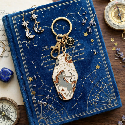 A gold-tone keychain with a Sagittarius-themed charm featuring a white and gold archer on a blue velvet book with gold constellation details, surrounded by celestial props including a compass, crystals, and dried lavender.