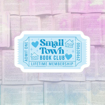 A die-cut sticker designed like a vintage boarding pass with the text 'Small Town Book Club' and 'LIFETIME MEMBERSHIP' in light blue on a white background, shown against a soft-focus background of open books.
