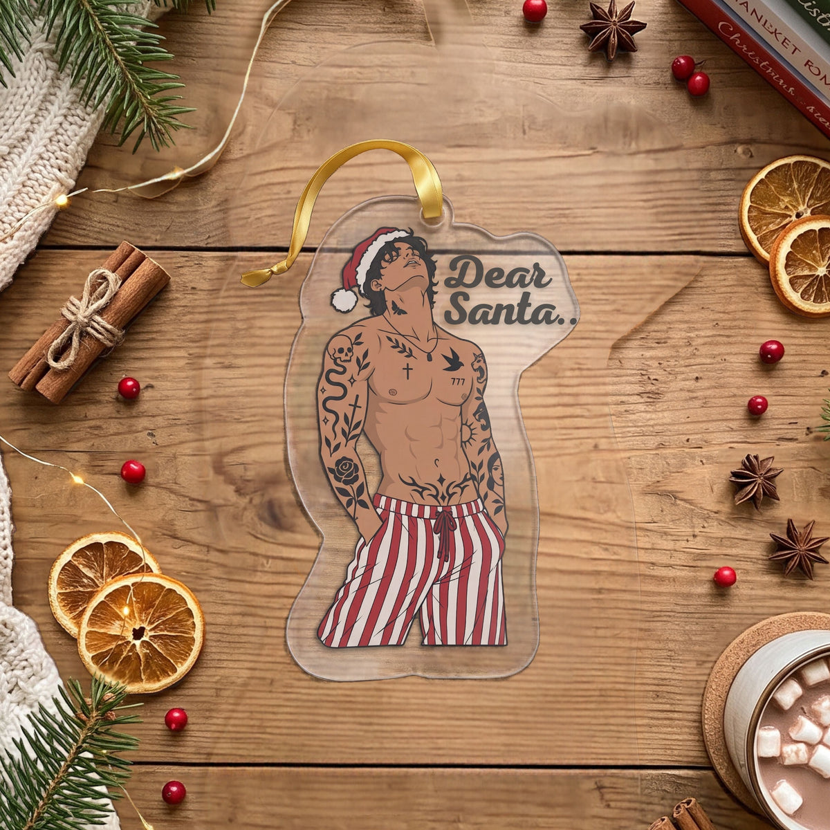 A Christmas ornament shaped like a shirt featuring a tattooed man in red-and-white striped pants and a Santa hat, with the text 'Dear Santa.' on a wooden tabletop surrounded by pine branches, dried orange slices, cinnamon sticks, a mug of hot chocolate with marshmallows, and stacked books.