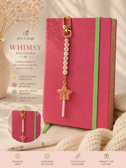 Pink notebook with a gold bag charm featuring letter beads spelling "Whimsey" pearls and a star on a beige background.