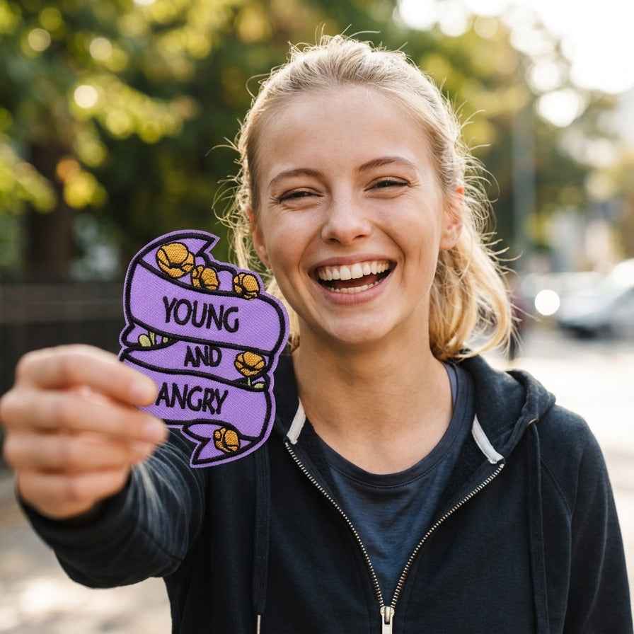 A smiling blonde woman outdoors holds up a purple sticker that reads 'YOUNG AND ANGRY' with cartoon angry faces.