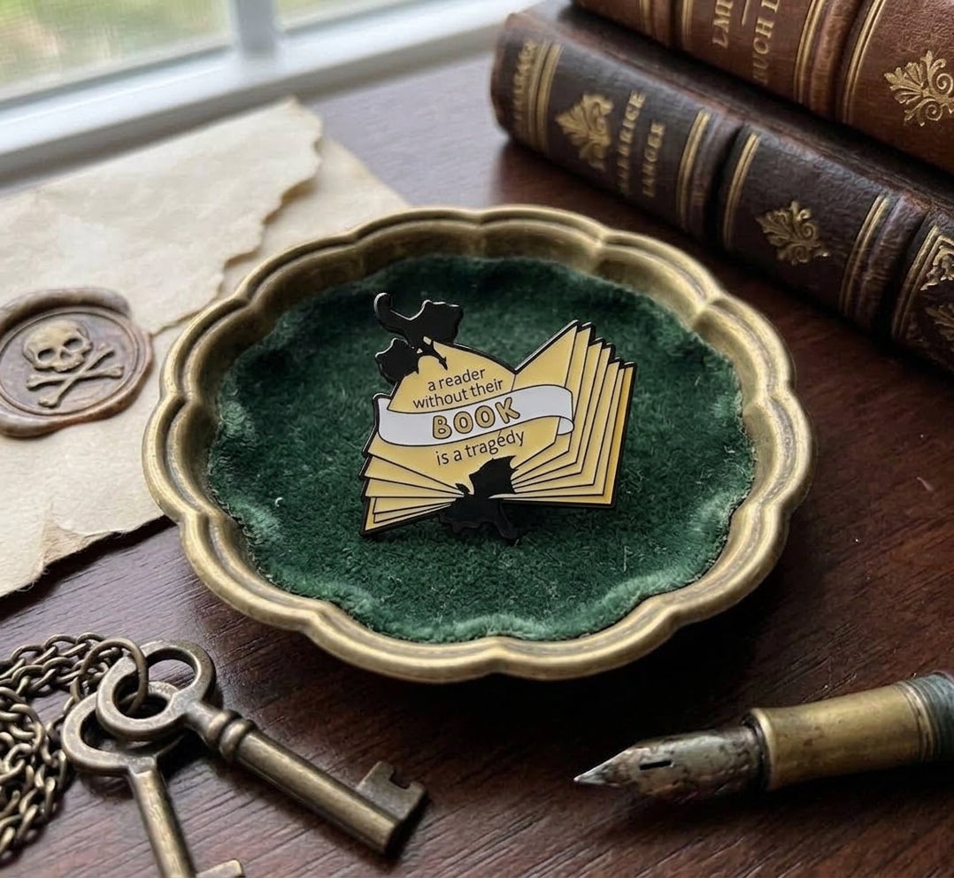 A book-themed enamel pin displayed on a green velvet insert inside a scalloped gold dish, styled on a wooden table with antique books, keys, parchment, and a fountain pen.
