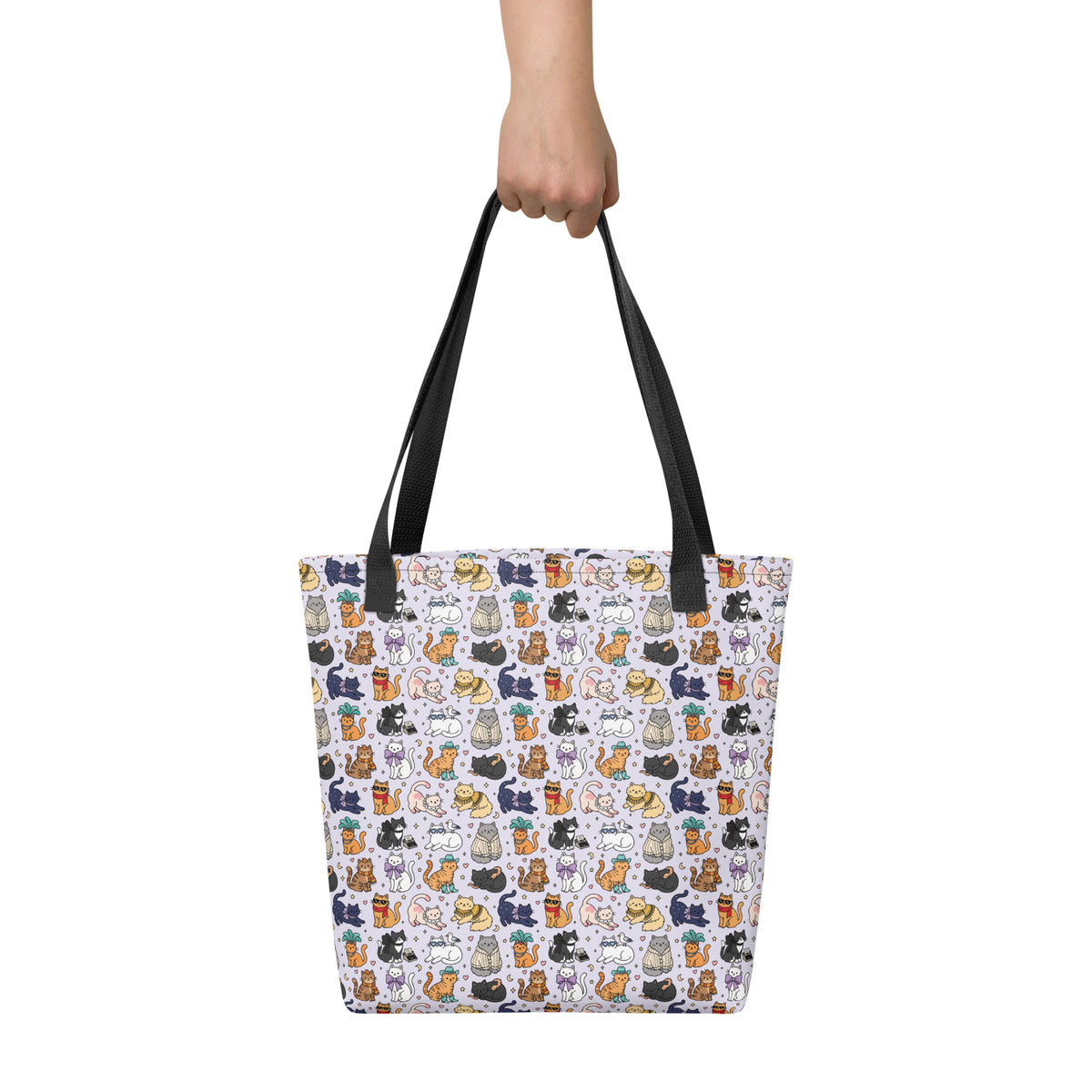 A tote bag with a white background and a colorful cartoon cat pattern, held by a hand with black shoulder straps, on a solid white background.