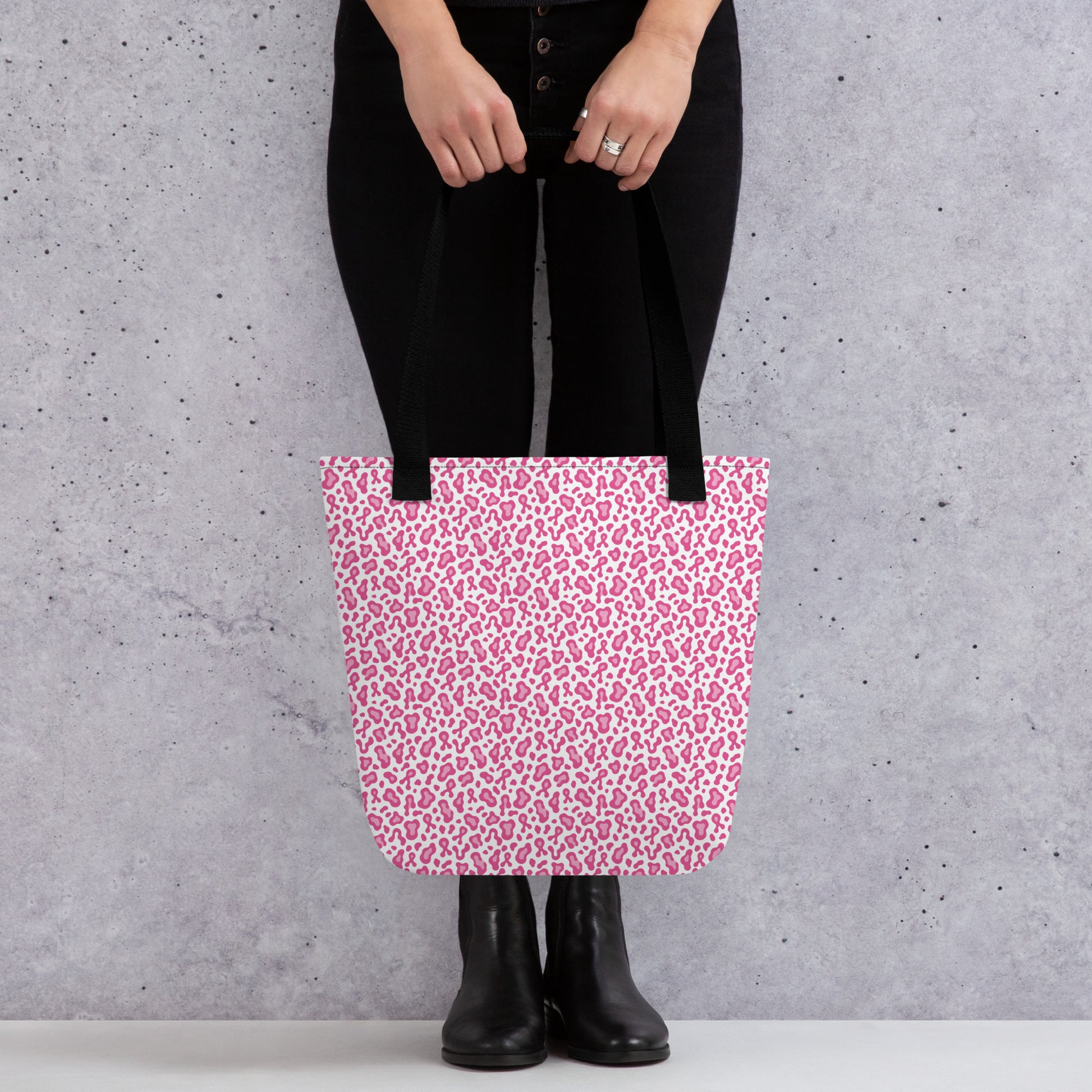 A person wearing black pants and black boots holds a pink and white leopard print tote bag against a textured gray concrete wall.