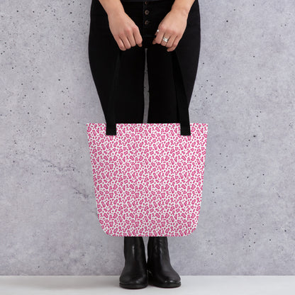 A person wearing black pants and black boots holds a pink and white leopard print tote bag against a textured gray concrete wall.