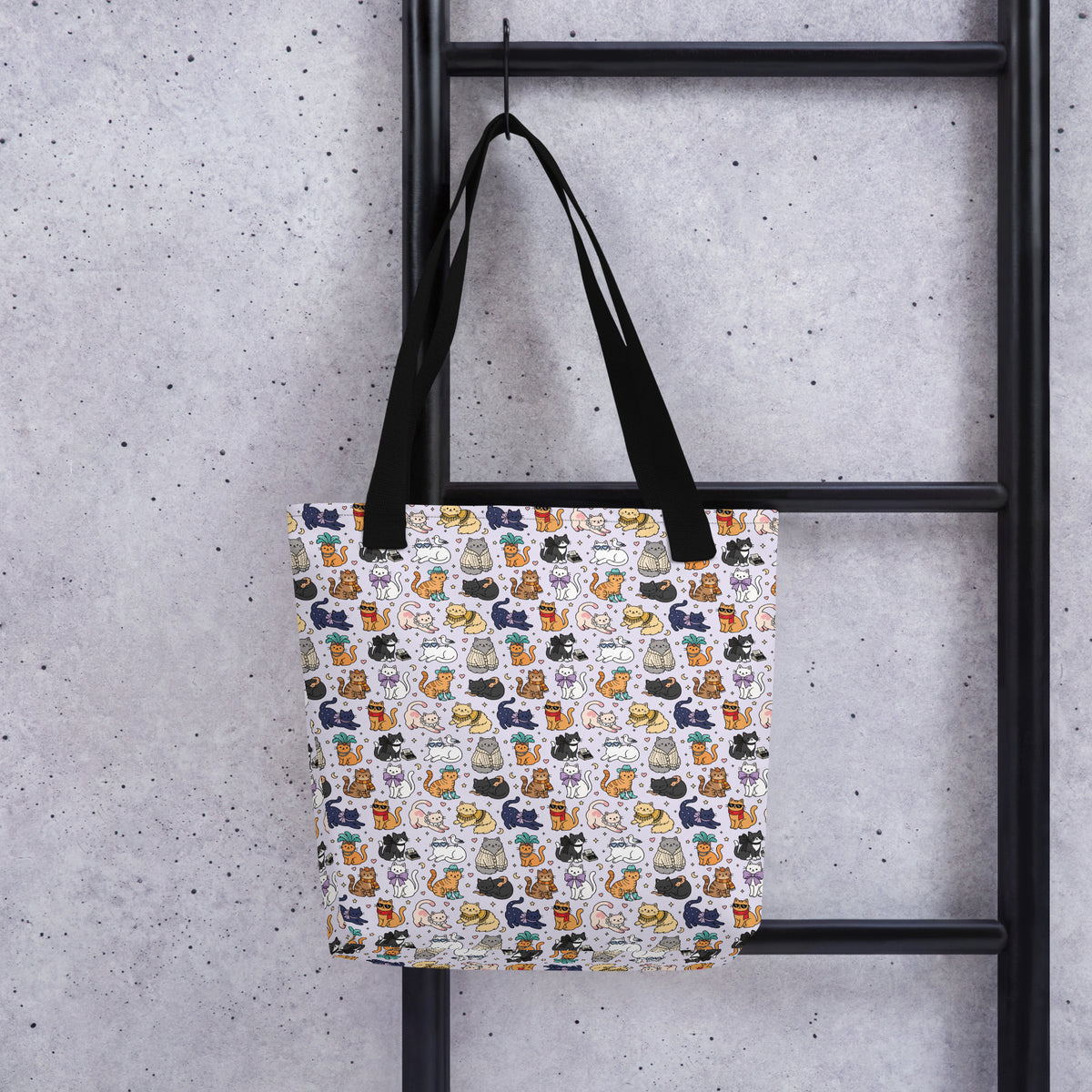 A tote bag with a white background and a colorful pattern of illustrated cats, hanging from a black metal ladder against a textured gray concrete wall.