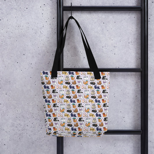 A tote bag with a white background and a colorful pattern of illustrated cats, hanging from a black metal ladder against a textured gray concrete wall.