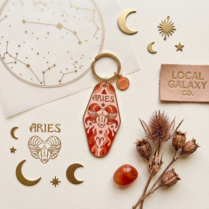 An Aries zodiac keychain with a red and orange ram illustration and the word 'ARIES' on a gold key ring, styled on a cream background with gold celestial cutouts, dried thistle stems, and a leather patch reading 'LOCAL GALAXY CO.'.