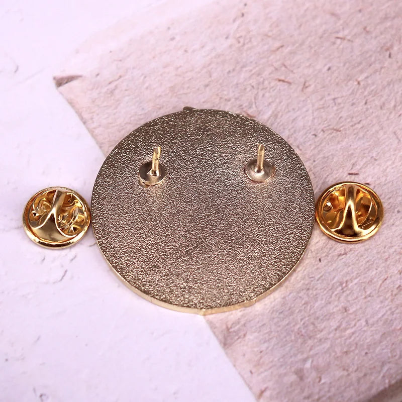 A round textured gold-tone pinback badge or medallion shown from the back, with two small gold post pieces visible, placed on a light pink textured surface with two matching gold pin-back pieces at the sides.