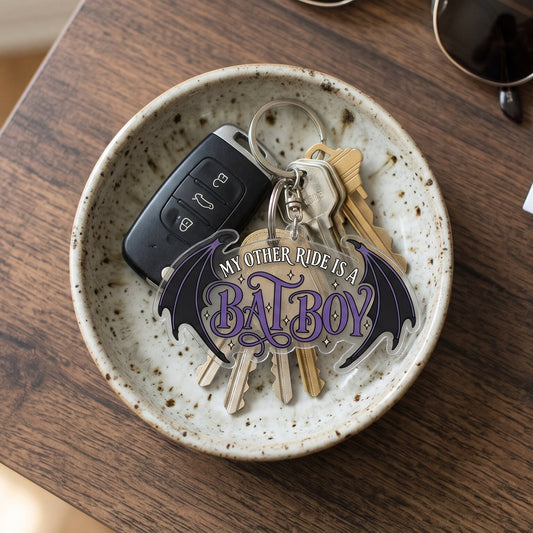 A 2-inch custom clear acrylic keychain with "My Other Ride Is A Bat Boy" written in purple font between dark bat wings, connected to a ring of house and car keys resting in a speckled ceramic bowl.