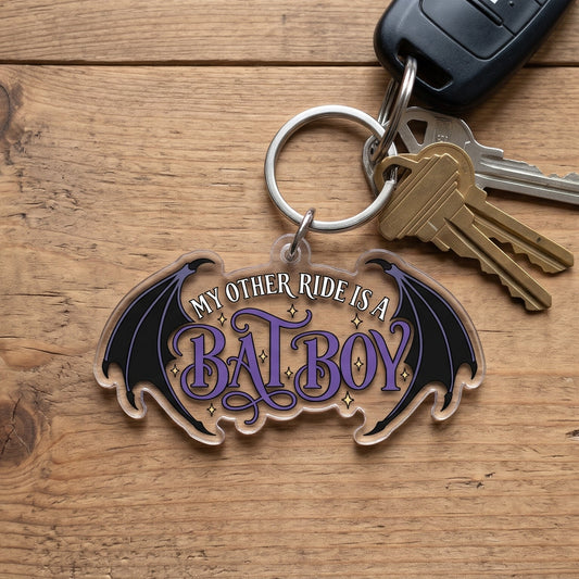 A 2-inch die-cut clear acrylic keychain featuring "My Other Ride Is A Bat Boy" with purple lettering and bat wings, attached to a keyring with car and house keys on a natural wood surface.