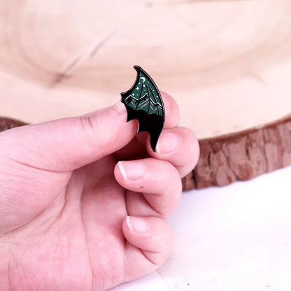 Hand holding a black and green bat wing-shaped pin against a wooden background