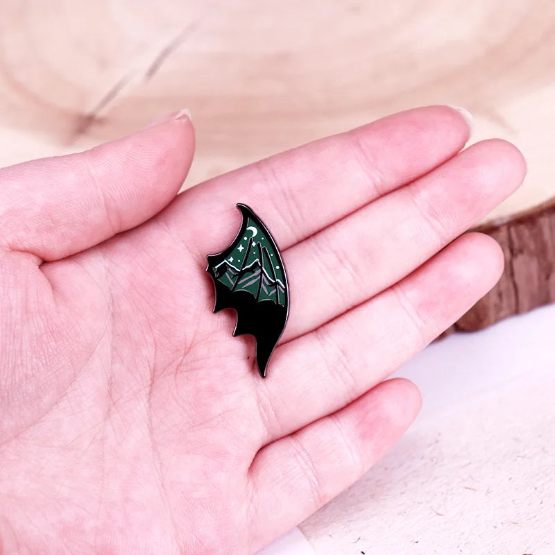 Black and green bat wing shaped pin with a mountain and starry sky design held in a hand against a neutral background
