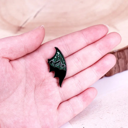 Black and green bat wing shaped pin with a mountain and starry sky design held in a hand against a neutral background
