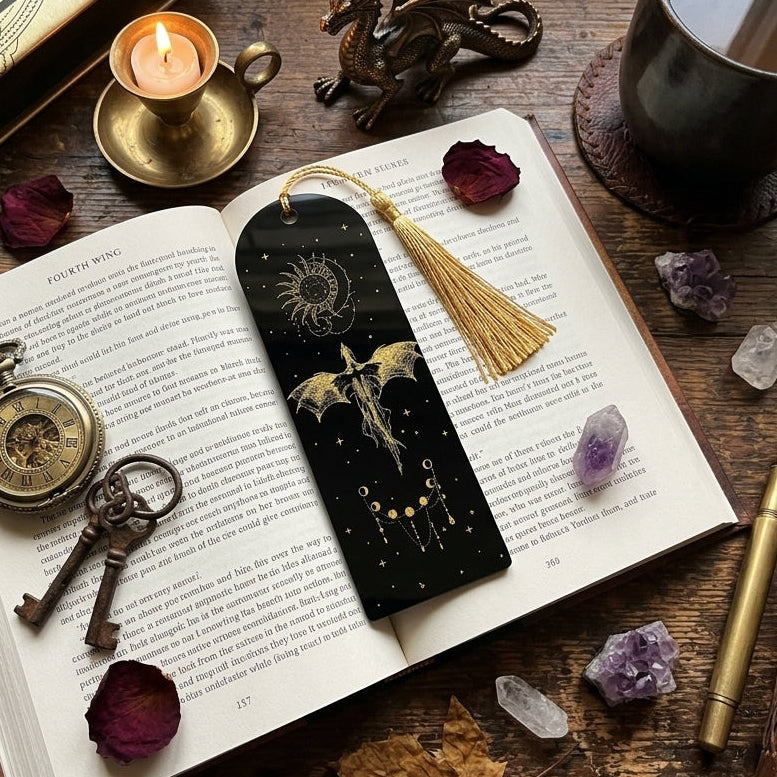 A black tassel bookmark with gold celestial and dragon illustrations rests on an open book surrounded by autumn-themed props including dried roses, crystals, a pocket watch, keys, a candle, and a coffee mug on a wooden table.