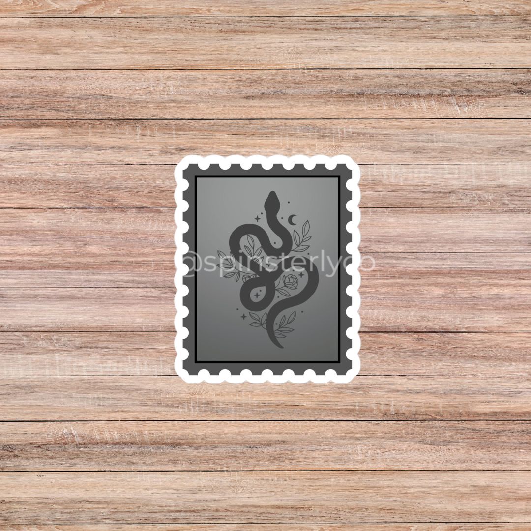 A rectangular die-cut sticker with a white border and scalloped edges, featuring a dark gray background with a black snake coiled over a rose and crescent moon. The sticker is displayed on a light brown wooden plank background.