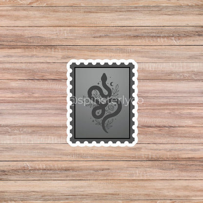A rectangular die-cut sticker with a white border and scalloped edges, featuring a dark gray background with a black snake coiled over a rose and crescent moon. The sticker is displayed on a light brown wooden plank background.