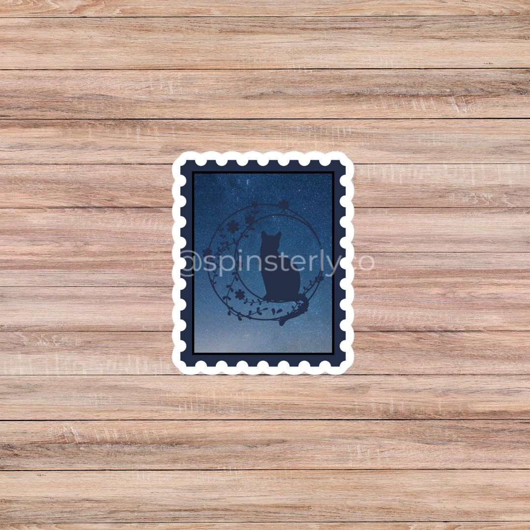A square die-cut sticker with a white scalloped border, featuring a dark blue night-sky design with a black cat silhouette and a circular wreath motif. The sticker is placed on a light brown wooden plank background.
