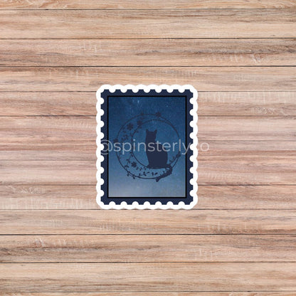 A square die-cut sticker with a white scalloped border, featuring a dark blue night-sky design with a black cat silhouette and a circular wreath motif. The sticker is placed on a light brown wooden plank background.