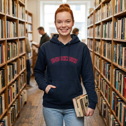 A smiling young woman stands in a library aisle wearing a navy hoodie with 'BOOKISH' printed in red across the chest, paired with light blue jeans and holding a book.