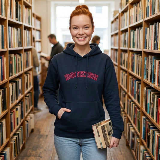 A smiling young woman stands in a library aisle wearing a navy hoodie with 'BOOKISH' printed in red across the chest, paired with light blue jeans and holding a book.