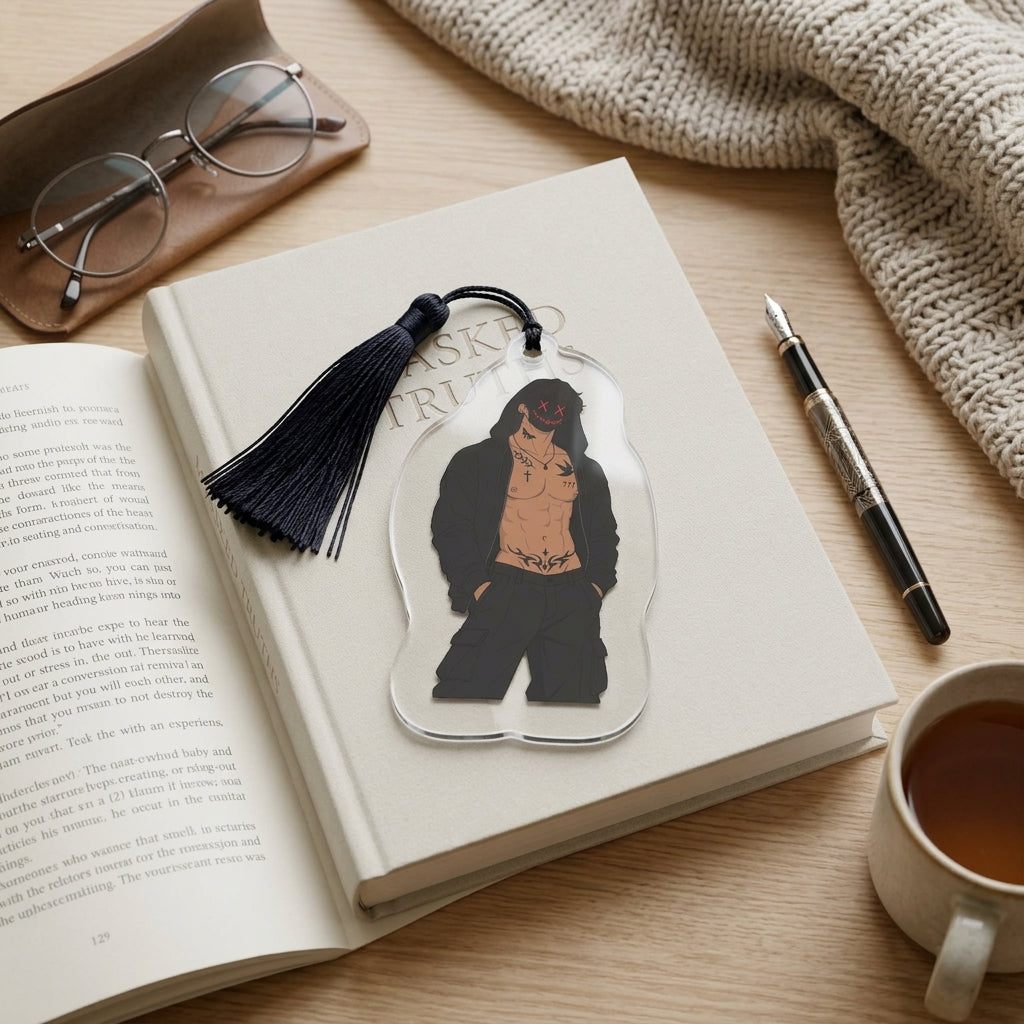 A clear acrylic bookmark featuring a black and tan illustration of a shirtless man with a red tattoo, resting on an open book. The bookmark has a black tassel and is styled on a wooden desk with glasses, a pen, a mug of tea, and a knitted blanket.
