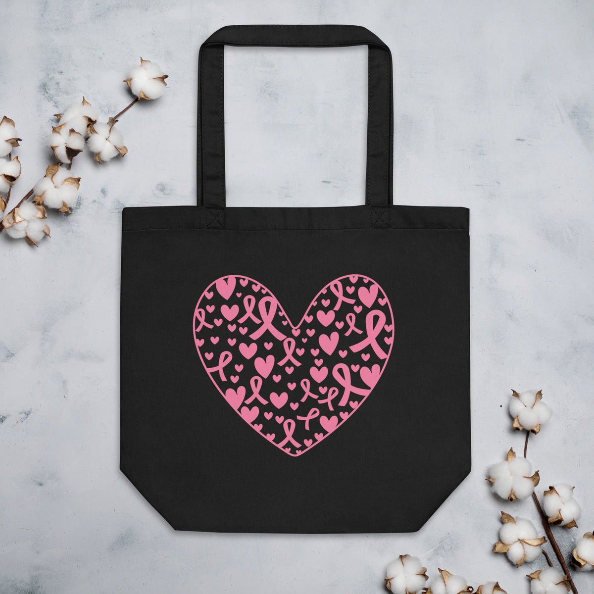 Black tote bag with a pink heart pattern made of breast cancer awareness ribbon and heart motifs, on a light gray surface with cotton branches.