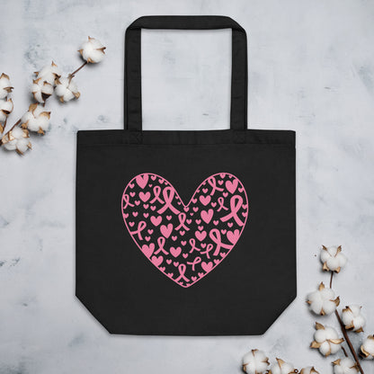 Black tote bag with a pink heart pattern made of breast cancer awareness ribbon and heart motifs, on a light gray surface with cotton branches.