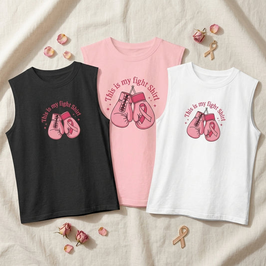 Three sleeveless tank top shirts in black, pink, and white, laid flat on a beige fabric background with pink rose petals and small awareness ribbon props. Each shirt has a pink boxing glove and pink awareness ribbon graphic with the text 'This is my fight shirt'.