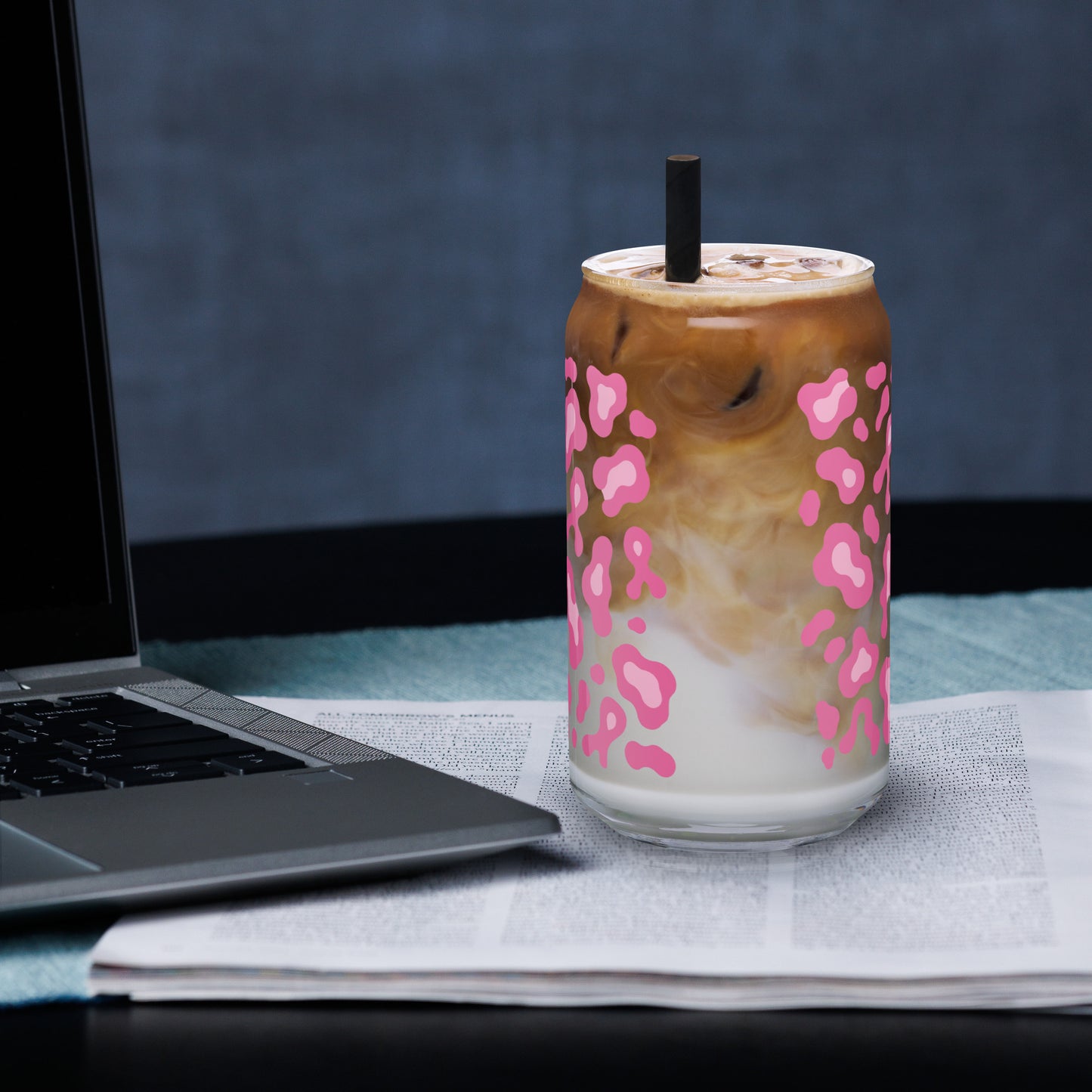 A clear glass can-shaped tumbler with a pink leopard print pattern, filled with an iced coffee drink and a black straw, set on a table next to an open laptop.