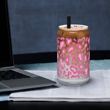 A clear glass can-shaped tumbler with a pink leopard print pattern, filled with an iced coffee drink and topped with whipped cream and a black straw, set on a desk next to an open laptop.